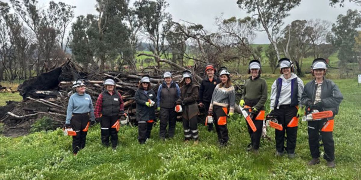 Eleven women in helmets, visors and chainsaw chaps standing with chainsaws in front of a storm-debris pile.